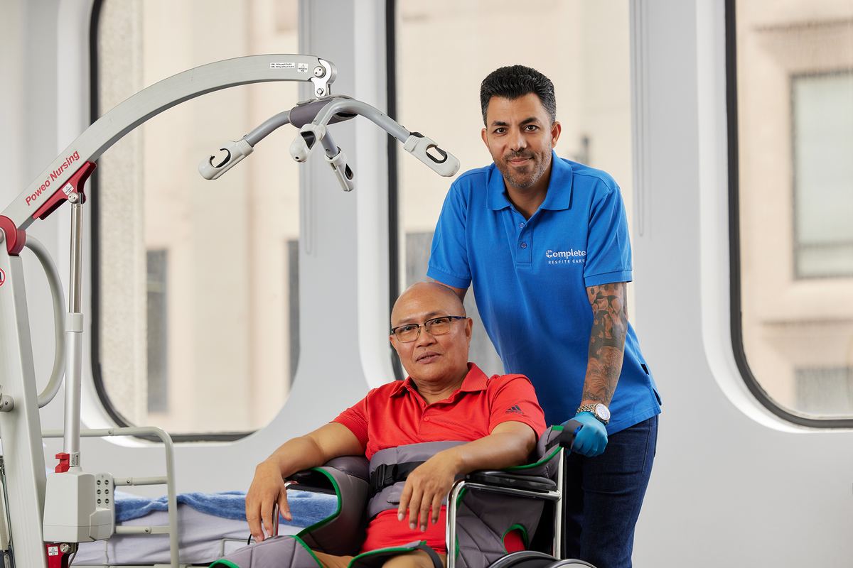 A carer standing with a client seated in a wheelchair beside a patient hoist