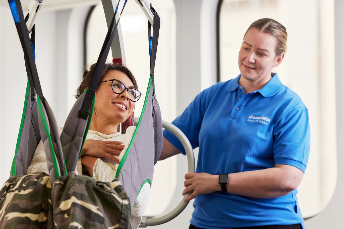 A smiling client seated securely in a lifting sling while a carer guides the hoist